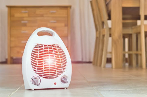 A white space heater, used in place of a broken furnace, with a red glowing light, placed on a tiled floor.