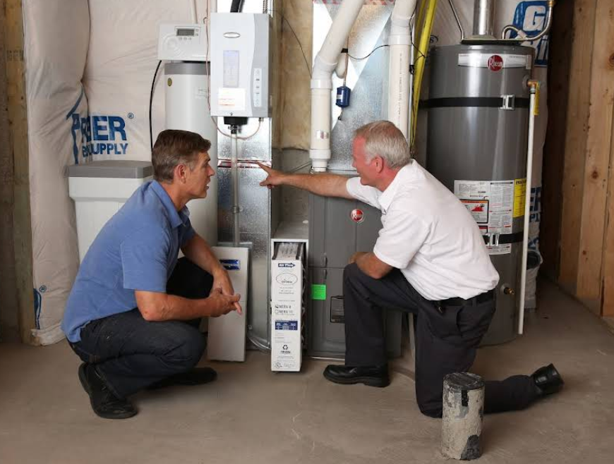 Technician kneeling and pointing at a furnace in a basement while discussing with a homeowner who is crouched down, listening.