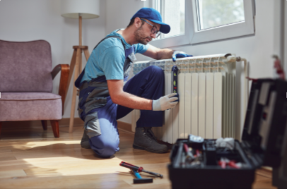 A male HVAC technician in a blue shirt, glasses, and a blue cap kneels on the floor next to a white radiator, using a level tool to check its alignment.